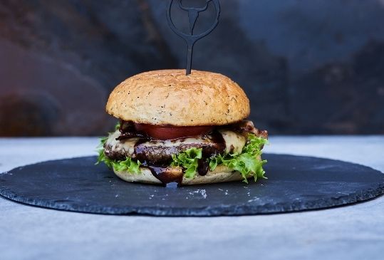 Saftiger Burger mit Patties von Fitmeat, Tomaten, Speck und Salat auf rundem Teller