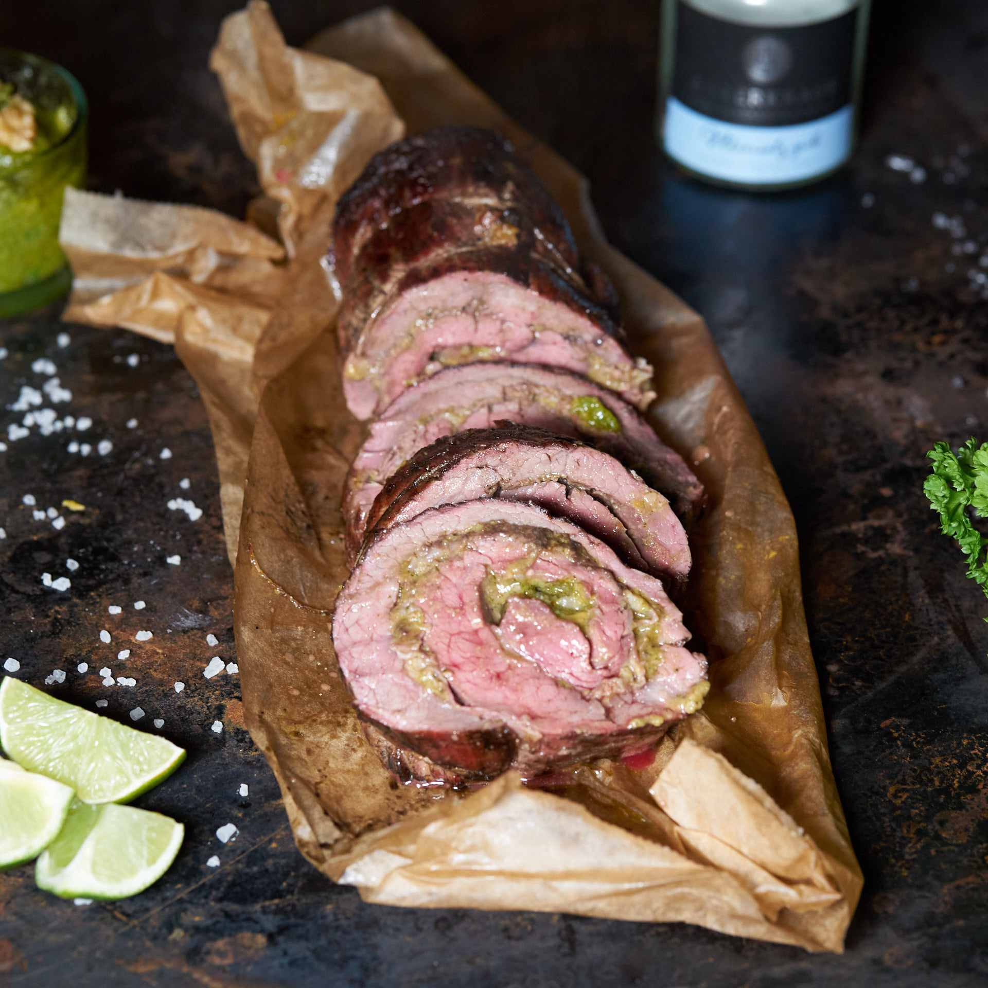 Gegrillte und mit Pesto gefuellte Flanksteak Rolle auf Backpapier mit Meersalz und frischen Limetten
